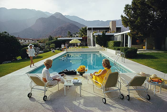 A desert house in Palm Springs designed by Richard Neutra for Edgar J. Kaufmann. Lita Baron (far left) approaches Helen Dzo Dzo (left) and Nelda Linsk, wife of art dealer Joseph Linsk. Original Artwork: A Wonderful Time - Slim Aarons.